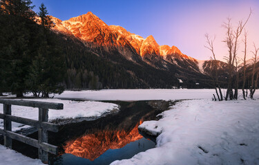 Snowcapped Alps mountains reflect warm colors in the partially frozen Anterselva lake (North Italy) at sunset in winter. Snow and ice in the foreground, tree and forest in the background.