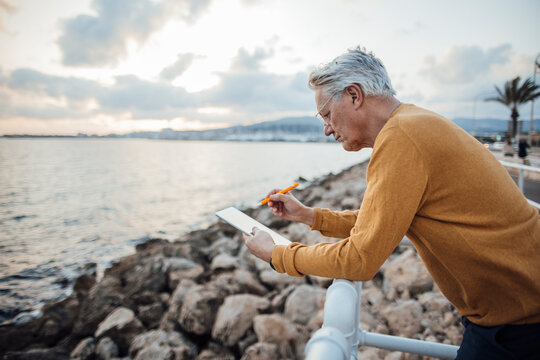 Senior man using tablet PC leaning on railing in front of sea