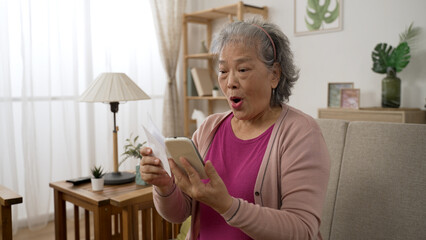 excited asian senior woman checking lottery numbers on mobile phone and cheering in surprise for winning prize in the living room at home.