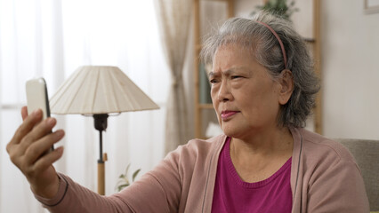 Asian old lady with presbyopia is holding her mobile phone far from eyes as she is having trouble reading prints on the screen clearly in the living room at home