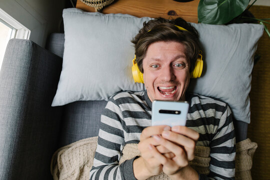 Excited Man Using Smart Phone And Wearing Headphones Lying On Bed At Home