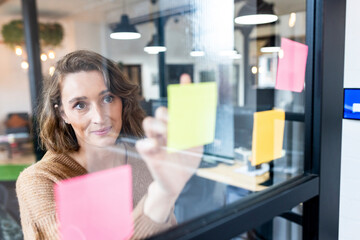 Smiling businesswoman sticking adhesive notes seen through glass