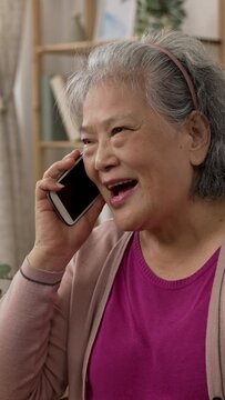 Vertical Screen: happy asian grandmother is smiling while enjoying chatting with friend on the mobile phone in a modern bright living room at home