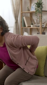 Vertical Screen: Asian grandma suffering chronic backache is dropping down in the couch and massaging her lower back in pain while trying to stand up in the living room at home