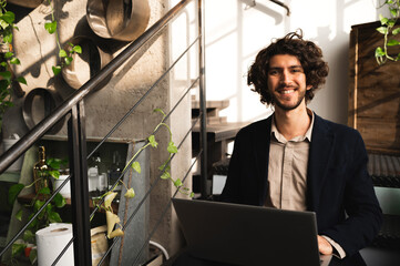 Hipster student posing at camera smiling. Guy sitting on a stair in a lounge and using computer laptop