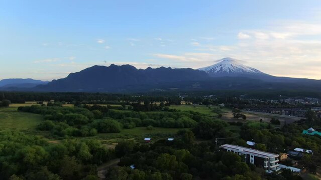 Beautiful aerial establishing shot of Pucon, Chile. Green trees and plants surround town in foreground with huge volcano, Villarrica Volcano in background.