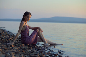 Beautiful young woman sitting on shore of lake in summer dress at sunset. Portrait of a romantic...