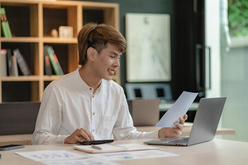 Serious young asian man working in office at desk. Holding and looking at document. Graph. Planning in office. calculation report Using laptop and calculator, online meeting concept