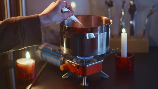 Portable stove usage at home during blackout, cooking on a tourist gas burner, stiring dish in a steel pot. Beauiful domestic background, burning candles. Surviving in Ukraine during war. HQ 4k