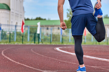 Fototapeta premium Long-legged athlete in a sports uniform prepares for the starting race, kneads the muscles of the legs. Runner kneads his legs in stadium before race on a circular treadmill with a rubber coating. 