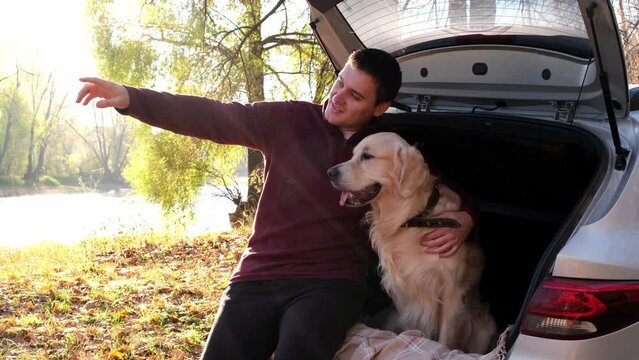 A happy man and his dog sit in the trunk of a car and cuddle. A young man with a golden retriever on a car trip in an autumn forest.