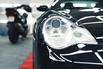 Sports car headlamp. Shiny black sports car in garage. Camera focus on the foreground. Blurred white motorcycle in the background. High quality photo
