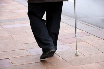 Legs of limping man in black trousers walking with a cane on city street. Concept for disability, old age, blind person