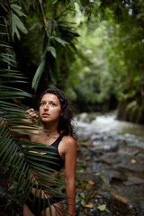 Portrait Young slender girl posing in the jungle hiding behind a palm leaf. Wildlife and a girl