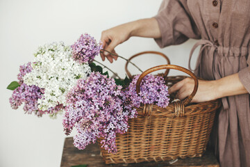 Stylish woman with beautiful lilac flowers in wicker basket in rustic room. Female in linen dress...