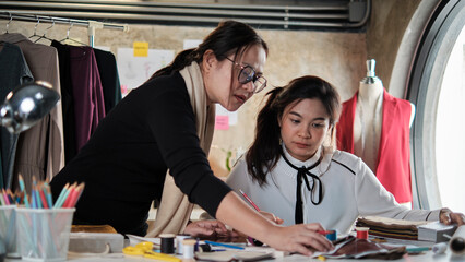Asian middle-aged female fashion designer teaches a young teen trainee tailor in studio with colorful thread and sewing fabric for dress design collection ideas, professional boutique small business.