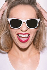 Beautiful woman close-up studio portrait with white sunglasses and red lipstick making joyful facial expression. Model wearing white shirt and have sun tan. Bright yellow filter applied. Toned image