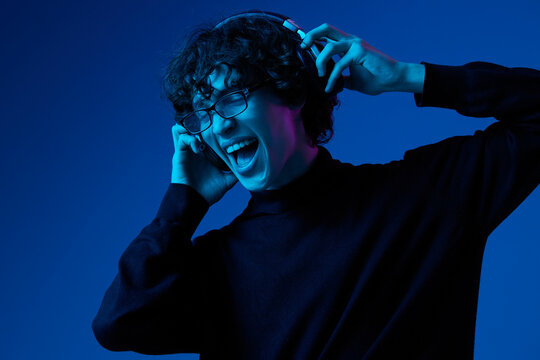 Teenage man wearing headphones listening to music and dancing and singing open mouth with glasses, hipster lifestyle, portrait blue background, neon light, style and trends, mixed light, copy space