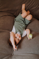 a happy little girl is lying at home on the couch eating a green apple