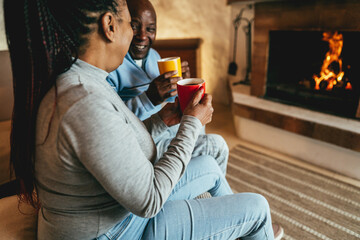 African couple drinking hot chocolate in front of cozy fireplace at home - Winter lifestyle concept...