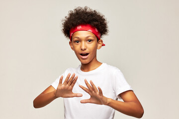 Portrait of funny afro kid boy doing no or stop gesture with palms refusing to play, smiling with opened mouth, wearing red bandana. Children, emotions and facial expressions. Body language