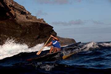 One man paddling an outrigger canoe next to big cliffs and crashing waves.