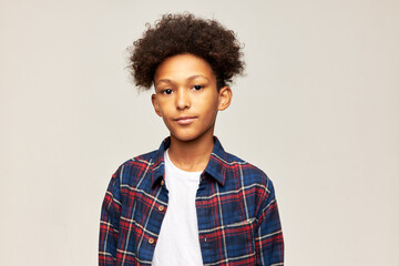 Studio image of smart serious teenage boy with afro hairstyle in plaid shirt posing in gray studio...