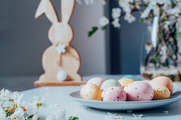 Wooden Easter bunny with blooming tree branches in vase and colored easter eggs i on the plate on the white kitchen table with flower petals. Happy Easter spring card. Selective focus. Copy space.
