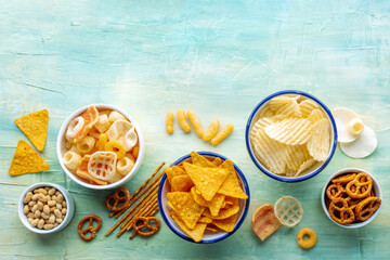Salty snacks with a place for text. Party food. Potato chips, nachos, crackers and other appetizers in bowls, shot from the top on a teal background