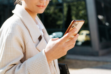 Close-up unrecognizable woman chatting, browsing internet on smartphone while standing in outdoor business district. Selective focus on mobile phone