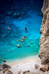 Tourists snorkeling in crystal clear water and anchoring boats next to a rugged rock face at Myzithres beach, Zakynthos, Greece