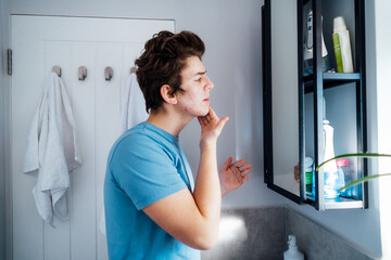 Concentrated caucasian teenage boy with acne problem take care his face skin at home. He looking in the mirror and cleaning face with washing lotion in bathroom. Teenager skin care process