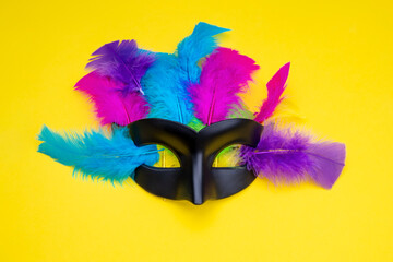 Venetian black carnival mask with colorful feathers on a yellow background.