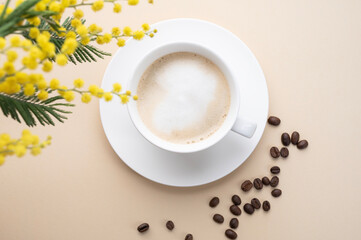 A spring bouquet with yellow mimosa flowers over a cup of coffee  cappuccino on a beige table with coffee beans. Concept of 8 March, happy women's day.