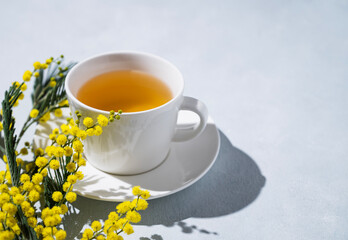 A spring bouquet with yellow mimosa flowers and a cup of herbal tea on a light background with morning shadow.