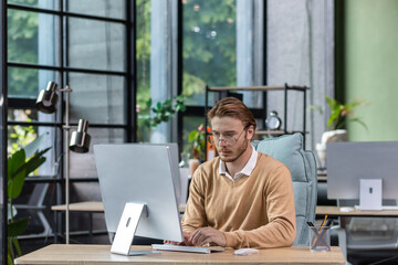 Serious and scorn-centered man in the office at work with a computer, man typing thoughtfully on keyboards, businessman young blond in sweater indoors loft, with green colors, programmer at work.