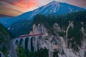 A local train of Rhaetian Railway coming out of the tunnel in a cliff crossing famous Landwasser Viaduct over a deep gorge with fall colors on the rocky mountainside in Filisur, Grisons, Switzerland
