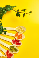 Flat lay composition with colored healthy vegan gummy bears  in eco wooden spoon with green leaves on yellow background, vitamins, healthy candies, top view, copy space.