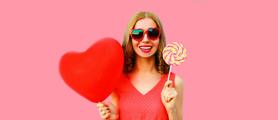 Portrait of happy smiling young woman with red heart shaped balloons and colorful lollipop having fun wearing sunglasses on pink background