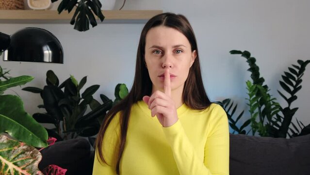 Portrait of suspicious young caucasian woman 20s wears yellow sweater looks at camera and brings index finger to mouth lips and she say shhh sitting alone on sofa at home. Shhh, woman secret finger