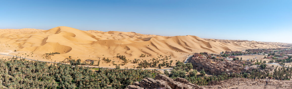 Panorama of Taghit town in Bechar, Algeria Sahara desert. Palm trees oasis, old ksor buildings and sand dunes with a blue clear sky. From Djebel Baroun mountain with unrecognizable tourists far away.