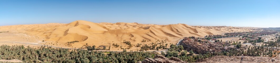 Panorama of Taghit town in Bechar, Algeria Sahara desert. Palm trees oasis, old ksor buildings and sand dunes with a blue clear sky. From Djebel Baroun mountain with unrecognizable tourists far away.