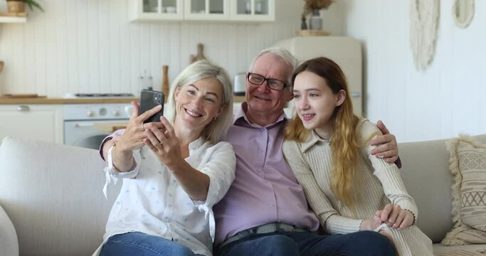 Happy multi generational family spend leisure rest on sofa staring at cellphone screen enjoy new application or take selfie. Grandpa his daughter and pretty teenage 12s granddaughter using modern tech