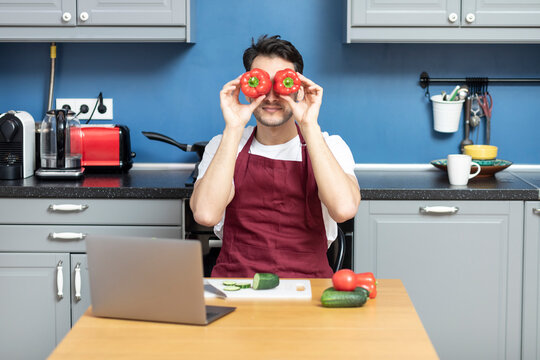 Young Funny Man Cooking In The Kitchen. He Watching Video Lessons About Cooking. Smiling Guy Wearing Red Apron Holding Red Peppers In Front Of His Eyes