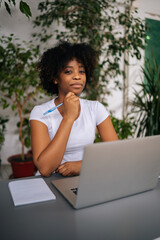 Vertical portrait of successful curly African American business woman holding pen in hand, serious looking at camera sitting at table in light home office room with modern biophilic interior design.