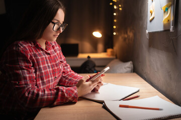 Cute teenage girl doing homework surfing internet on smartphone sitting at table. Portrait of focused student using mobile phone studying in dormitory room.