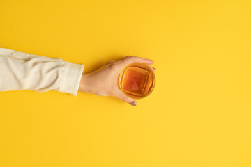 Hand reaching for a glass of alcohol. Overhead view of glass whiskey on yellow background