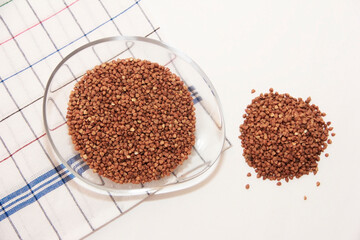 Small grains of natural brown buckwheat in a transparent plate on a light background.