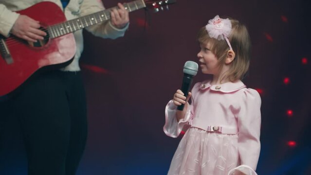 Little girl on stage in vintage dress, she sings into microphone and dances, her father plays acoustic guitar. Color music is shining. Performance on stage. Early development of children