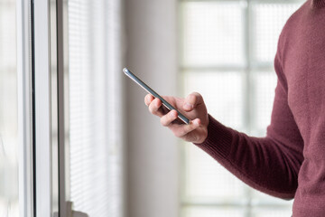 Close up of a man in sweater standing in front of windows at home and holding smartphone. Man hands holding smartphone